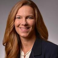 A smiling woman with long brown hair in a dark blazer is set against a gray background, reflecting her passion for digital healthcare at medbridge.