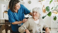 A caregiver in blue scrubs talks to an older woman in a wheelchair in a bright kitchen, showing medbridge hybrid care.