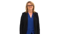 A woman in glasses, a black blazer, and a blue blouse stands against a white background.