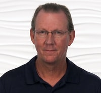 A middle-aged person with glasses and tied-back hair in a dark shirt stands against a white background, reflecting digital healthcare themes relevant to medbridge.