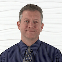 A man in a blue striped shirt and patterned tie smiles slightly against a white textured wall.