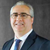 A gray-haired individual in a suit and tie smiles warmly, embodying digital healthcare expertise, against a dark background.