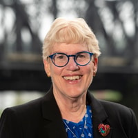 A smiling person with short light hair and blue glasses wears a black blazer with a heart-shaped pin and stands in front of a blurred bridge for medbridge.