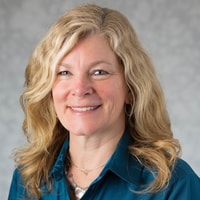 Smiling woman with wavy blonde hair, teal blouse, and necklace against a light gray background for medbridge healthcare solutions.