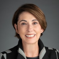 A smiling person with short brown hair wears a patterned black and white jacket, set against a gray background in the medbridge photo.