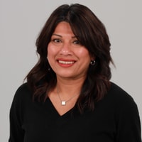 A person with dark hair smiles, wearing a black top and jewelry, against a neutral background.