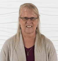 A blonde woman with glasses and a necklace stands against a textured white background, wearing a beige sweater over a purple top.