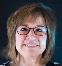 Individual with glasses and light brown hair smiling in a dimly lit setting.