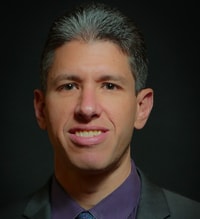 A man with short gray hair in a suit and tie, set against a dark background, looks at the camera for medbridge.