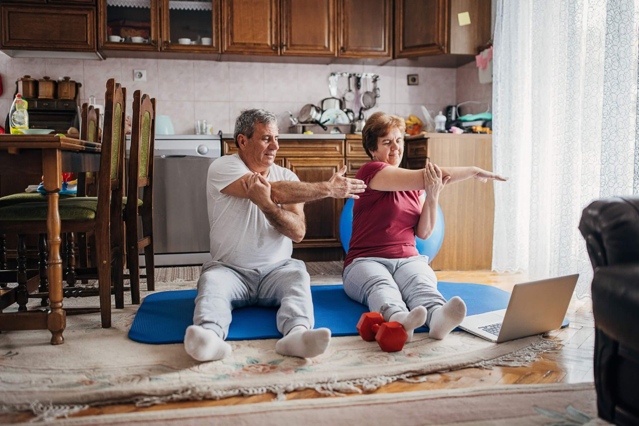 An elderly couple stretches on exercise mats in front of a laptop, likely watching a medbridge workout video.