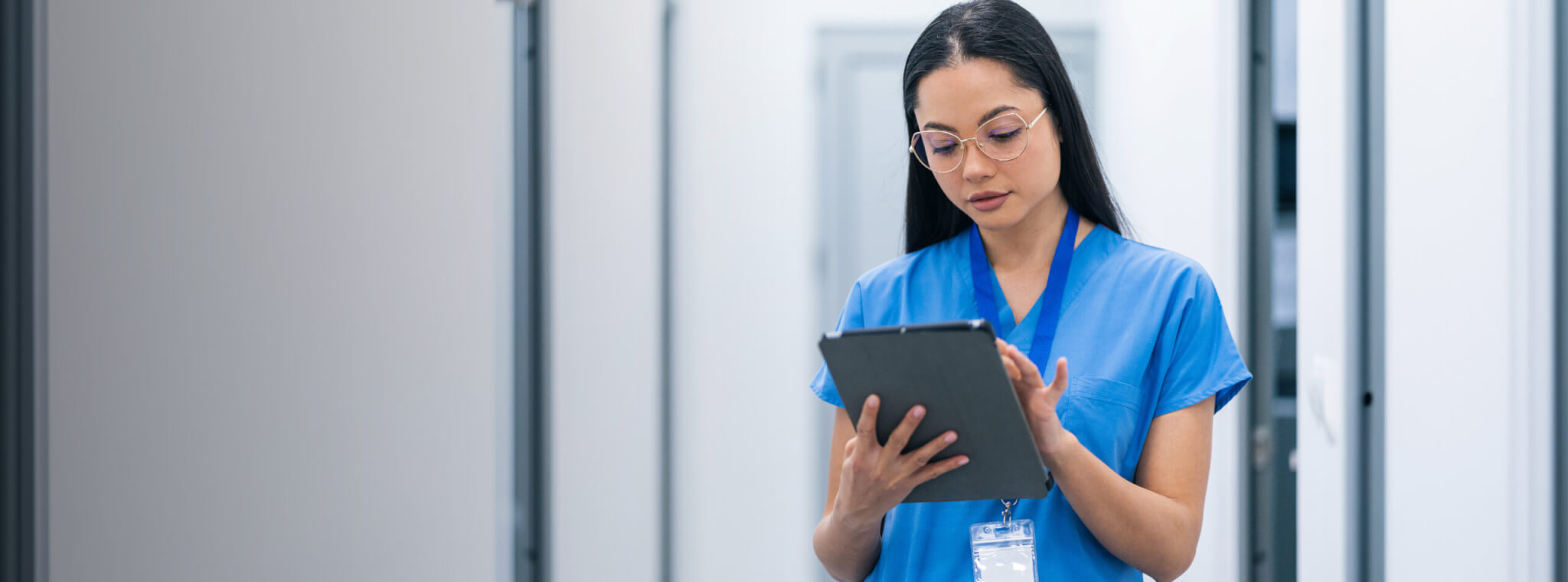 A healthcare professional in blue scrubs holds a tablet in a corridor.