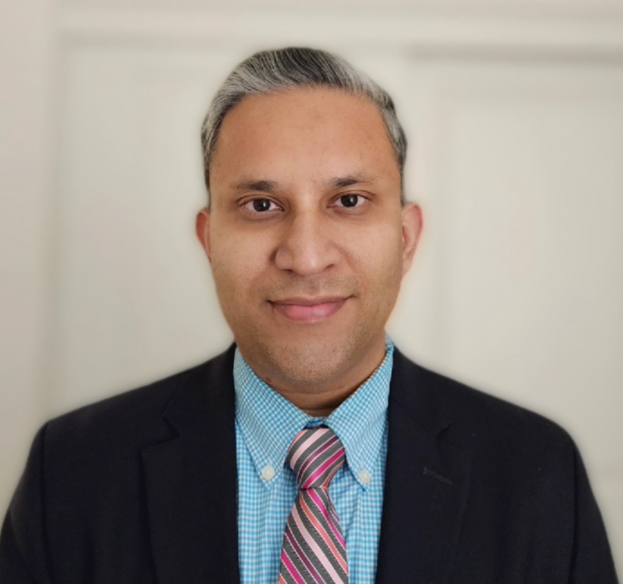 A man with short gray hair in a blue checkered shirt, pink tie, and dark blazer poses before a light background for medbridge.