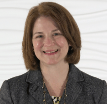 A woman with shoulder-length brown hair smiles, wearing a blazer and patterned top, against a textured wall, representing Medbridge's innovation in digital healthcare.
