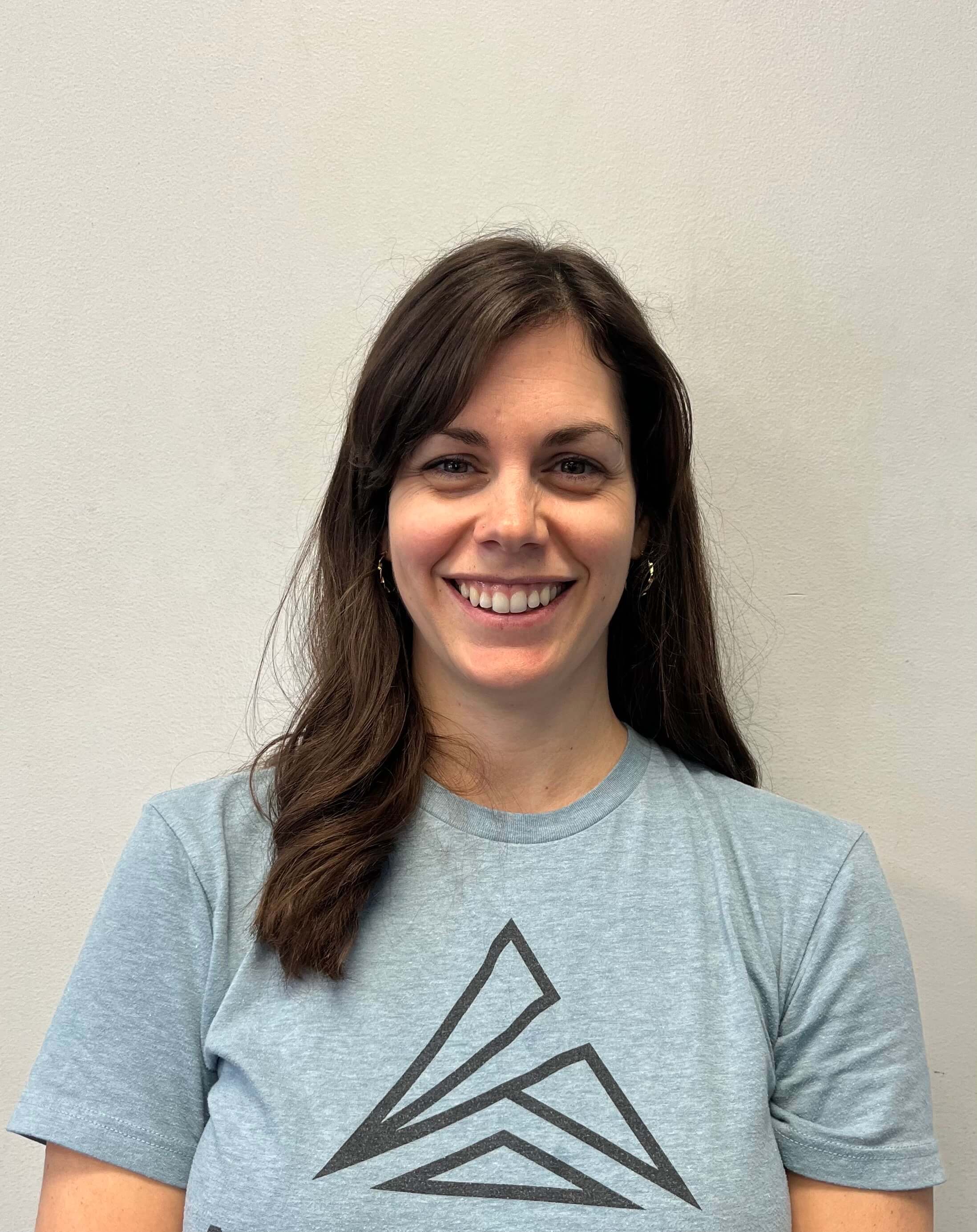 Smiling woman with long brown hair in a light blue t-shirt stands in front of a plain wall, reflecting medbridge professionalism.