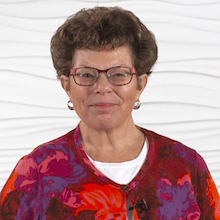A woman with short curly hair and glasses smiles warmly, embodying her passion for healthcare education, against a textured white background.