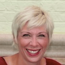 A smiling person with short blonde hair wearing a red outfit stands against a light brick wall.