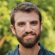 A bearded man stands outdoors with a slight smile, set against a backdrop of blurred greenery, symbolizing medbridge hybrid care's tranquility.