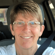 Person with short hair, glasses, and earrings, smiling in a car while wearing a white top.