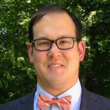 Individual in glasses and a suit with an orange-striped bow tie standing before greenery.