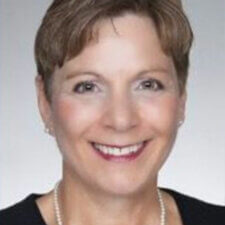A smiling person in a black top and pearl necklace stands against a gray background, embodying medbridge's blend of style and digital healthcare innovation.