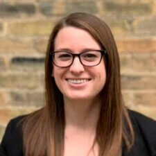 A confident woman with glasses and long brown hair smiles warmly against a brick wall, embodying passion for digital healthcare innovations at Medbridge.