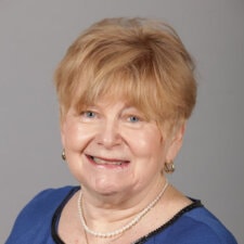 An older individual with light hair smiles warmly, wearing a blue top and pearl necklace.