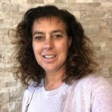 A smiling woman with curly brown hair stands in front of a stone wall, representing the charm of digital healthcare innovations at medbridge.