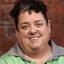 A person with short curly hair, wearing a green jacket, smiles warmly in front of a brick wall, capturing the spirit of healthcare education associated with medbridge.