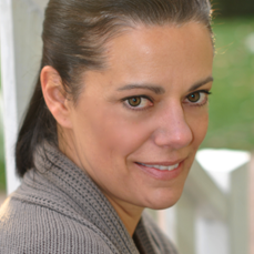 A dark-haired person in a gray sweater smiles slightly at the camera with a blurred outdoor backdrop.