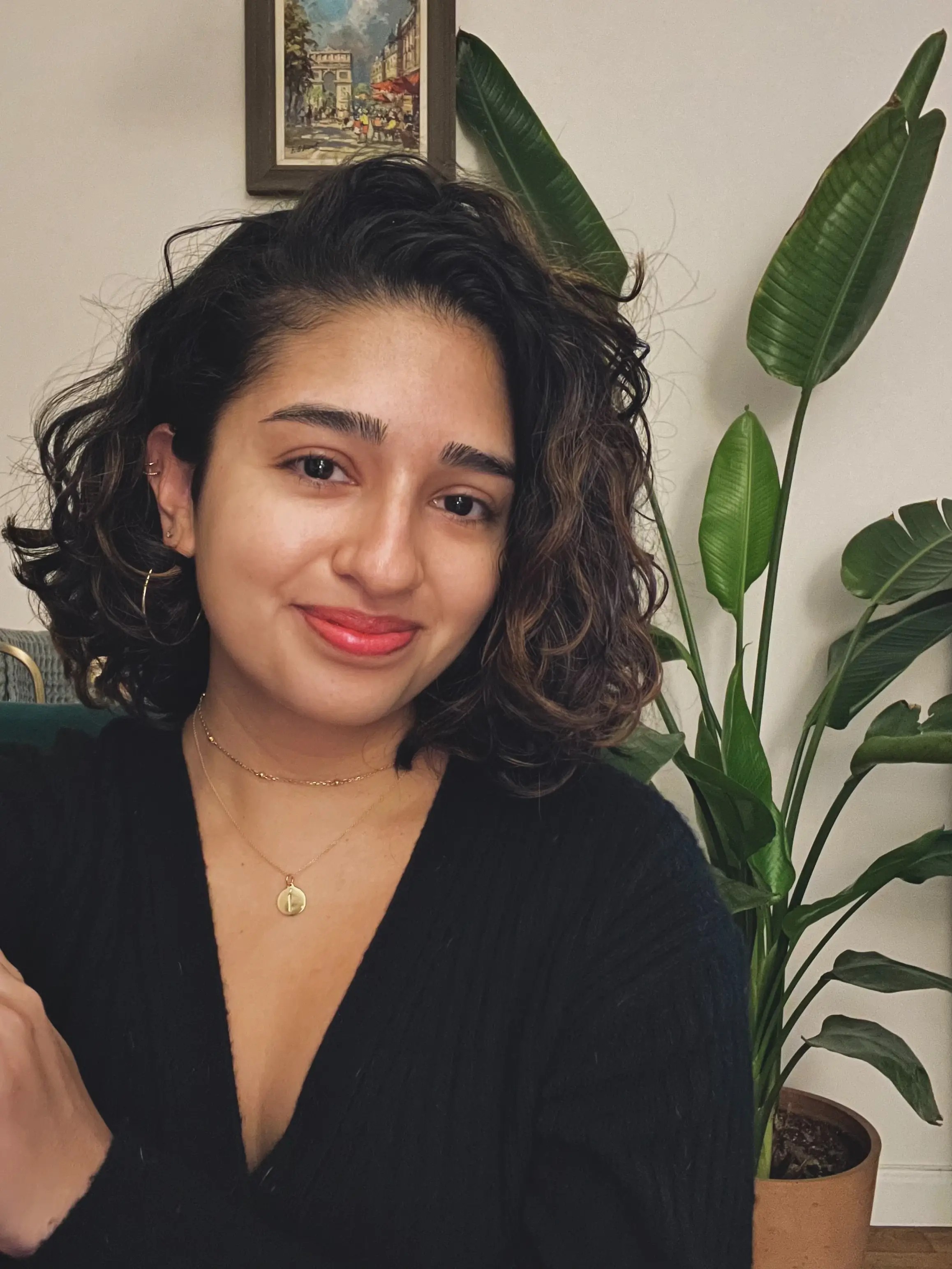 A person with curly brown hair and a black top smiles in front of a green plant and small framed art on a white wall.