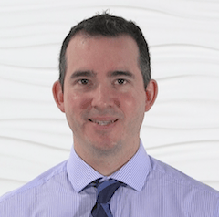 A person in a light purple shirt and dark blue tie stands against a white textured background.