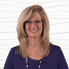 A smiling woman with blonde hair and glasses, wearing a purple top and necklace, stands in front of a white textured background, representing Medbridge's hybrid care.