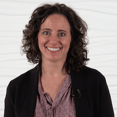A curly-haired person in a dark jacket and patterned blouse smiles warmly against a textured white background, embodying medbridge's vibrant spirit of healthcare education.