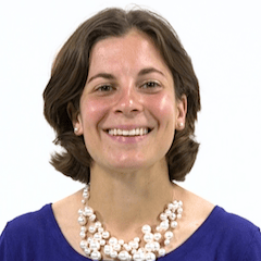 A smiling person with brown hair, wearing a blue top and pearl necklace, stands against a white background.