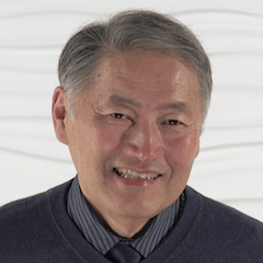 An older man with gray hair smiles in front of a textured white background, wearing a dark sweater over a striped shirt and tie.