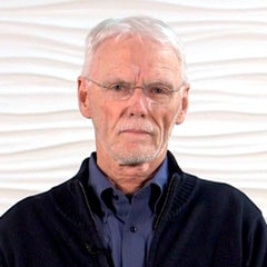 An older man with white hair and glasses in a dark sweater, representing healthcare wisdom, stands against a textured white background—perfect for medbridge.