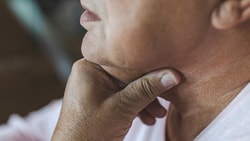 A person in a light shirt touches their throat, looking pensive or worried.