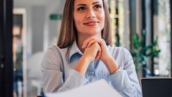 In a bright office, someone at a desk smiles with hands clasped, looking at another person.