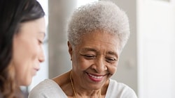 Two women, one younger and one older with short gray hair, chat and smile indoors in a casual or professional setting.