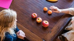 Two people sit at a wooden table with apples, tangerines, and a pink notebook.