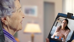 Elderly woman video chats on a tablet wearing a purple scarf in a cozy room using medbridge.