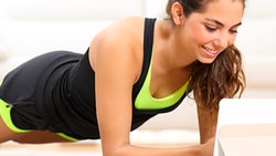 Woman planking in workout gear, watching a tablet, couch behind her in the room.