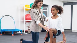 A therapist helps a girl with a knee exercise amid colorful therapy equipment.