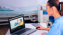 A healthcare professional in scrubs sits at a desk using medbridge on a laptop for a video call in a clinical setting.