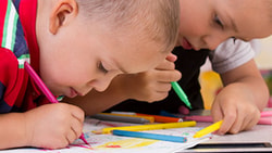 Two children color with markers at a table filled with art supplies.