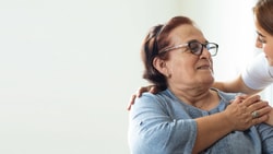 A caregiver in a white uniform converses with an older woman in a wheelchair against a plain background.