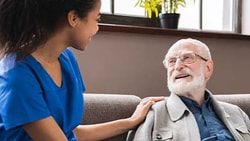 A Medbridge nurse in blue scrubs converses with an elderly man on a sofa.