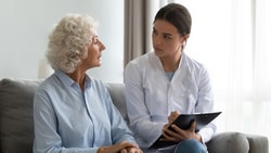 An elderly woman converses with a healthcare professional in a bright living room, with a wheelchair nearby.