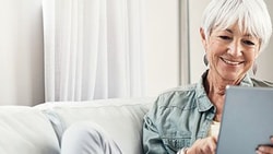 Smiling older woman with white hair uses a tablet on a softly lit couch, wearing a light shirt and earrings.