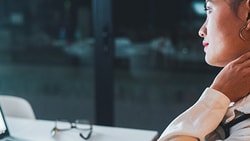 A woman in a white coat with a stethoscope massages her neck while looking at a medbridge screen.